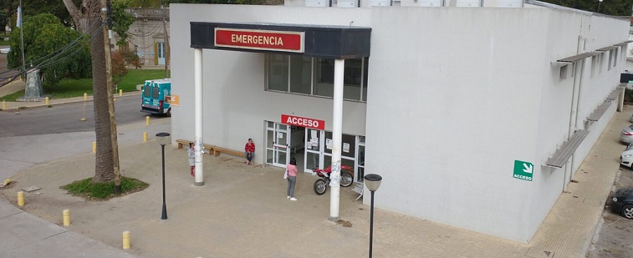 Se vé una parte del edificio del hospital Que recibió la donación de equipos informáticos. Es la entrada por la parte de emergencia. Tiene un cartel rojo que indica el acceso. una moto y tres mujeres se ven en la puerta de ingreso.