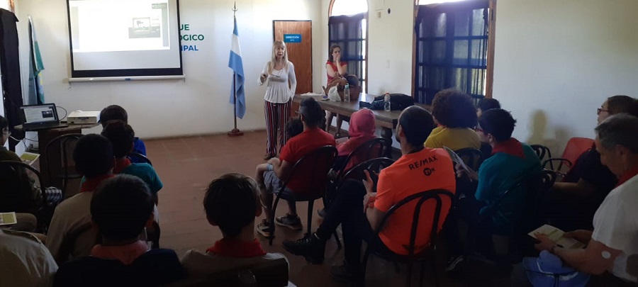 En un espacio interior del Parque Ecológico se ve parada a la Directora de EKOA brindar la charla. Sobre la pared e el fondo se vé una pizarra blanca y una bandera argentina de pie. La gente se encuentra sentada en sillas de espaldas escuchando a la expositora.