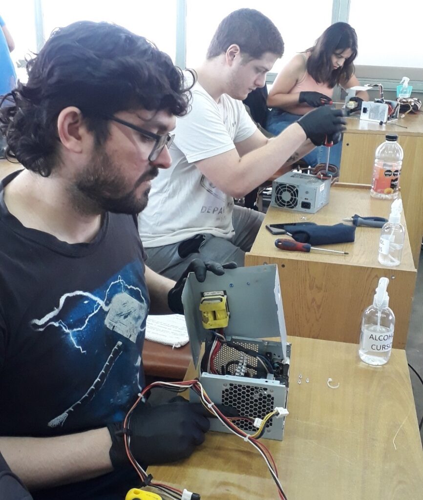 Tres personas sentadas en escritorios individuales realizando reparaciones o mantenimiento de fuentes de poder para computadora. La persona en primer plano, con gafas, camiseta negra con un diseño de martillo y rayos, y guantes negros, sostiene una fuente de poder abierta mientras la observa. La persona del medio, con camiseta blanca y guantes negros, usa un destornillador para trabajar en otra fuente de poder. Al fondo, una mujer con camiseta sin mangas color durazno y guantes negros también manipula una fuente de poder con un destornillador. Sobre los escritorios hay herramientas, botellas de alcohol en gel con etiquetas visibles, y otros objetos relacionados con la reparación. La escena está iluminada por luz natural que entra por ventanas grandes detrás de ellos.