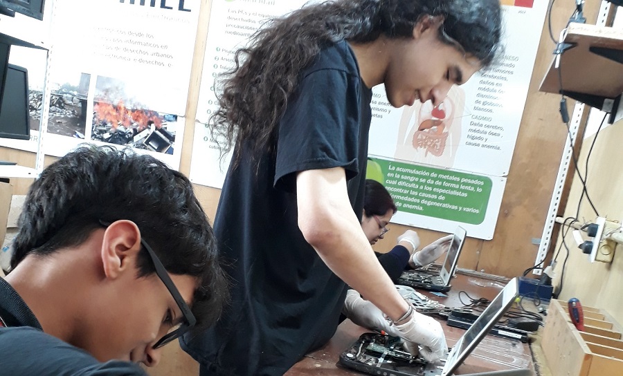 Estudiantes trabajando en la reparación o desmontaje de computadoras portátiles en un taller técnico. Ambos usan guantes para manipular los componentes electrónicos. La imagen muestra un ambiente de aprendizaje práctico con carteles informativos en la pared al fondo, relacionados con el manejo responsable de residuos electrónicos. Los estudiantes están concentrados en su tarea en un espacio equipado para la formación profesional.