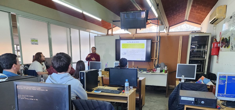 Aula del Programa EKOA con estudiantes sentados en bancos de frente un profesor. Se ve una pantalla con una presentación, computadoras, un televisor y una mesa
