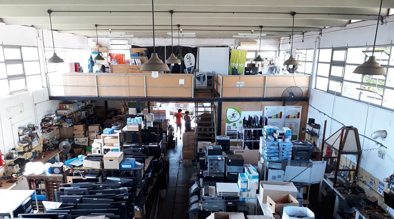 Interior de un gran almacén o depósito con estanterías y cajas llenas de equipos electrónicos, principalmente monitores, computadoras y otros dispositivos tecnológicos. Se observan dos personas de pie al fondo, cerca de la entrada, conversando. El espacio es amplio, luminoso por las ventanas laterales y tiene una mezzanina usada como oficina o espacio de trabajo.