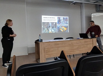 Curso del Programa EKOA Dos jóvenes trabajan en una mesa de laboratorio con componentes de computadora y herramientas. Uno de ellos manipula una fuente de alimentación, mientras el otro gesticula y organiza cables. En la mesa hay una botella de agua, dos laptops y diversos cables; fondo con más equipos y personas concentradas en tareas técnicas.