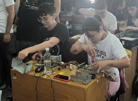Visita de Pablo Villena a EKOA.Tres personas sentadas alrededor de una mesa de reuniones observando una presentación en pantalla. En primer plano, a la izquierda, un hombre con camiseta negra; en el centro, una mujer con cabello rubio y otro hombre con cabello oscuro hablando. Tras la mesa, una pantalla muestra una diapositiva; la sala tiene ventanales grandes y estanterías con materiales.