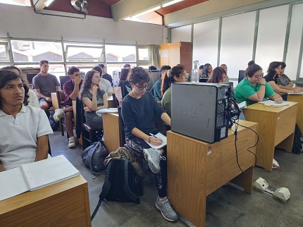 Visita de Pablo Villena a EKOA.Tres personas sentadas alrededor de una mesa de reuniones observando una presentación en pantalla. En primer plano, a la izquierda, un hombre con camiseta negra; en el centro, una mujer con cabello rubio y otro hombre con cabello oscuro hablando. Tras la mesa, una pantalla muestra una diapositiva; la sala tiene ventanales grandes y estanterías con materiales.